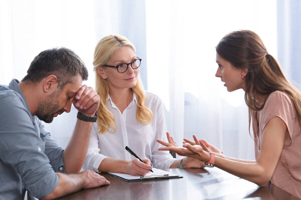 woman gesturing by hands and quarreling with stressed husband sitting with hand on forehead at table on therapy session by female counselor in office 
