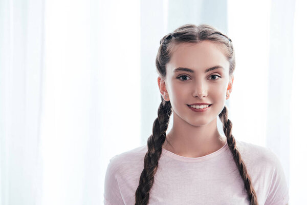 portrait of smiling teenage girl with plaits looking at camera in front of curtains at home