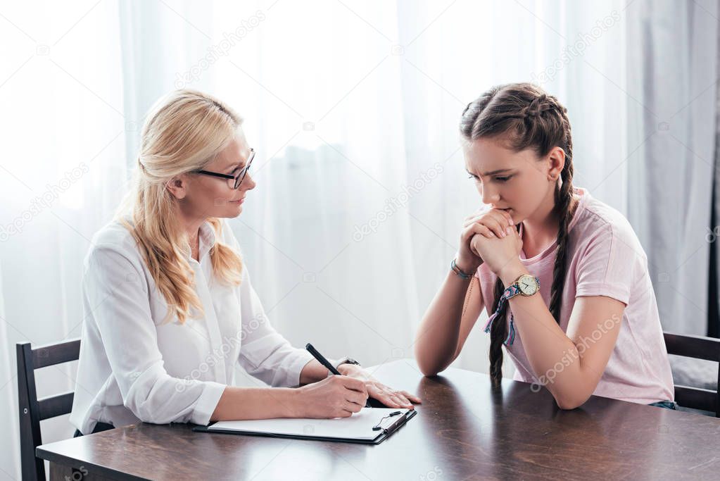stressed teenage girl sitting at table on therapy session by female counselor writing in clipboard in office 
