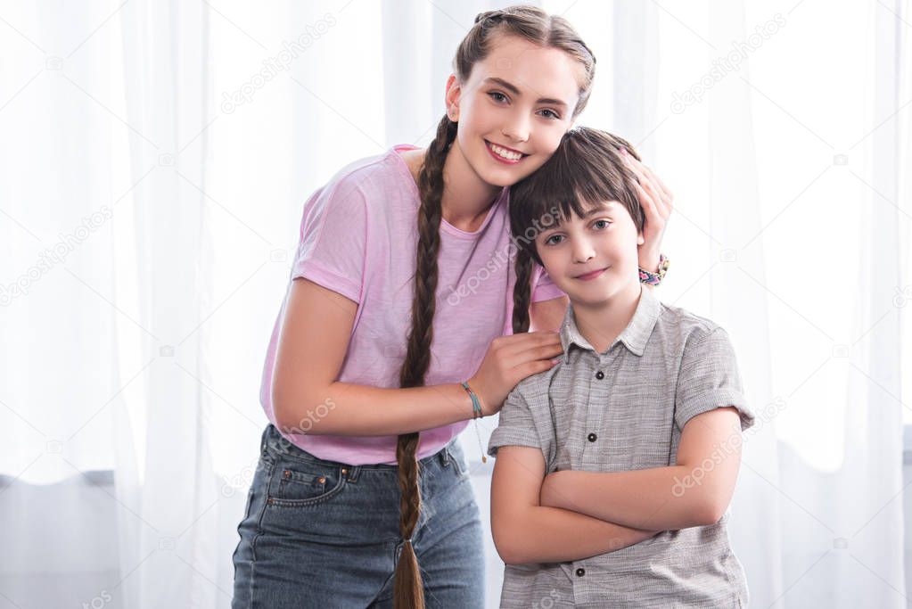 smiling teenage girl embracing little brother with crossed hands at home