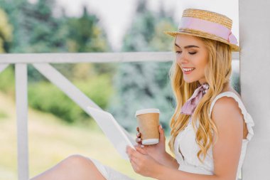 side view of beautiful smiling blonde girl in wicker hat holding coffee to go and using digital tablet clipart