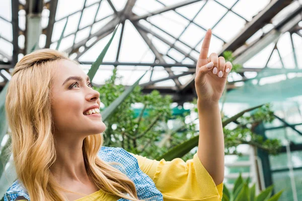 Hermosa Mujer Rubia Sonriente Apuntando Hacia Arriba Con Dedo Invernadero —  Fotos de Stock