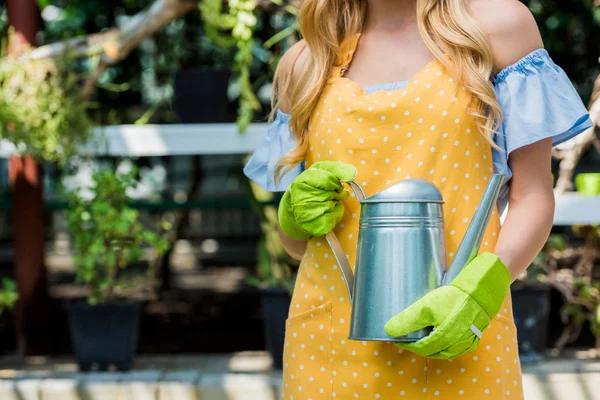 cropped shot of young woman in rubber gloves holding watering can in greenhouse