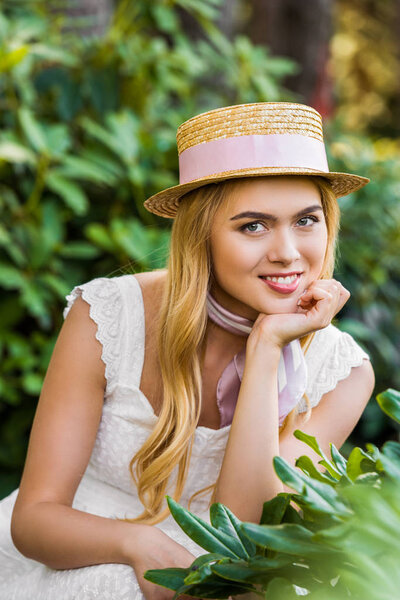 portrait of beautiful young blonde woman in wicker hat with ribbon smiling at camera between green plants
