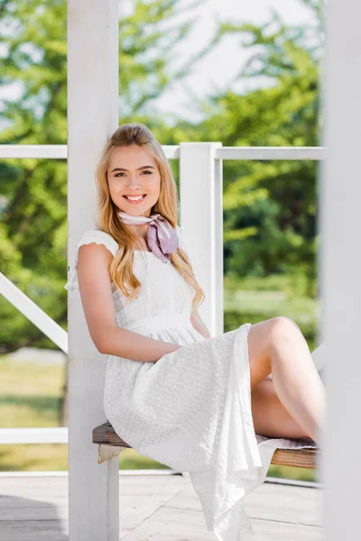 bankta oturan ve kameraya gülümseyen beyaz elbiseli sarışın güzel genç kadın