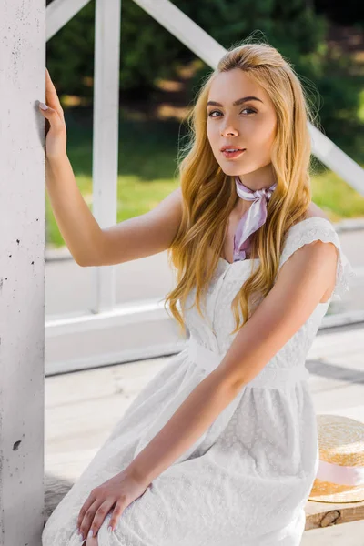 bankta oturan ve kameraya gülümseyen beyaz elbiseli sarışın güzel genç kadın