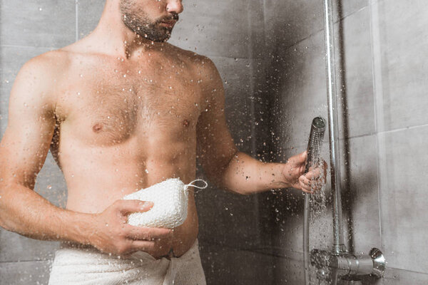 Handsome bearded man holding sponge behind shower glass