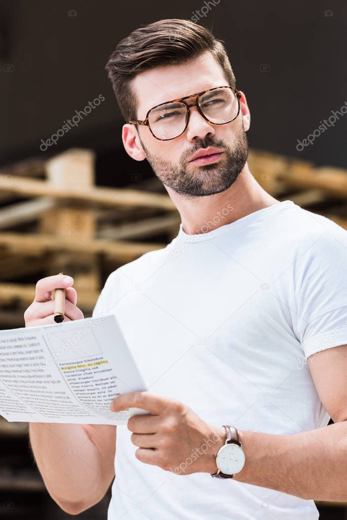 Fashionable confident businessman reading business document and holding cigar by wooden palettes