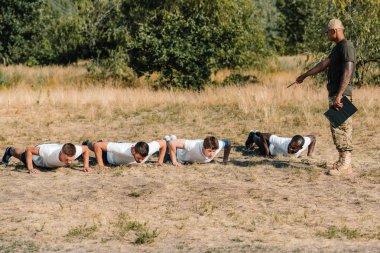 tactical instructor with notepad examining multicultural soldiers doing push ups on range clipart