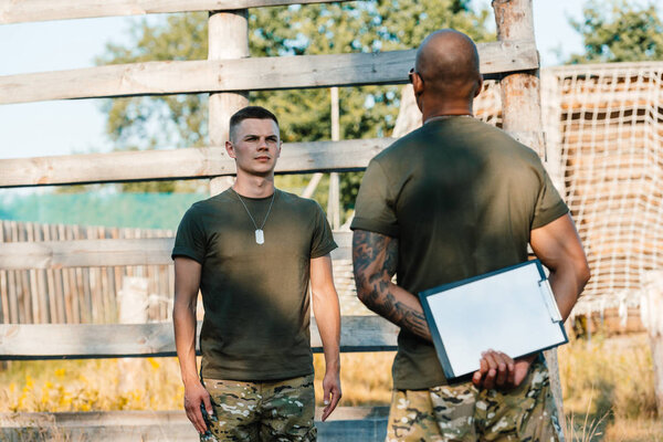 african american tactical instructor with notepad and young soldier in military uniform on range