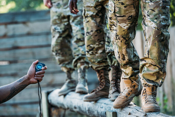 partial view of tactical instructor with stop watch examining multiracial soldiers during obstacle run on range