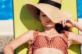close-up portrait of attractive young woman in straw hat and bikini relaxing on sun lounger at poolside