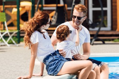 Beyaz t-shirt ve güneş gözlüğü havuz üzerinde birlikte oturan genç aile gülümseyen