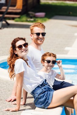 güzel genç aile beyaz t-shirt ve havuz üzerinde birlikte oturan ve kameraya bakarak güneş gözlüğü