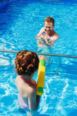 father and little daughter playing with water guns in swimming pool clipart
