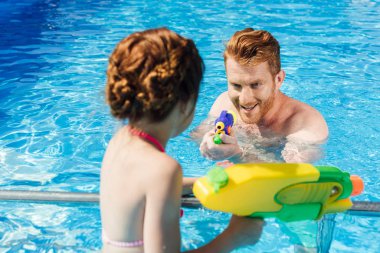 young father and daughter playing with water guns in swimming pool clipart