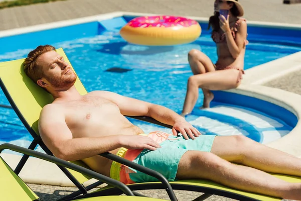 handsome young man tanning on sun lounger while his girlfriend sitting on poolside