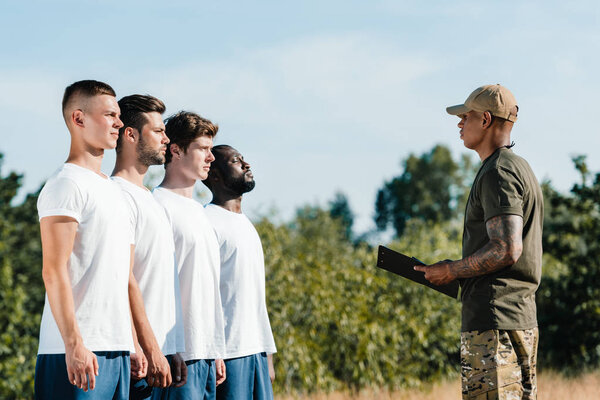 side view of african american tactical instructor with notepad and multiracial young soldiers on range