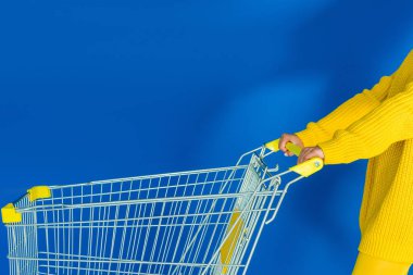 Cropped view of female hand on shopping cart on blue background clipart