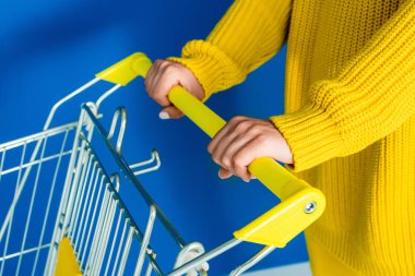 Cropped view of woman in yellow clothes holding shopping cart on blue background clipart