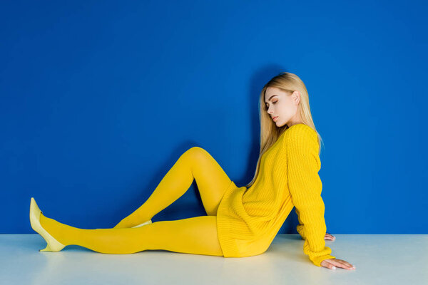 Elegant blonde woman dressed in yellow sitting on floor on blue background