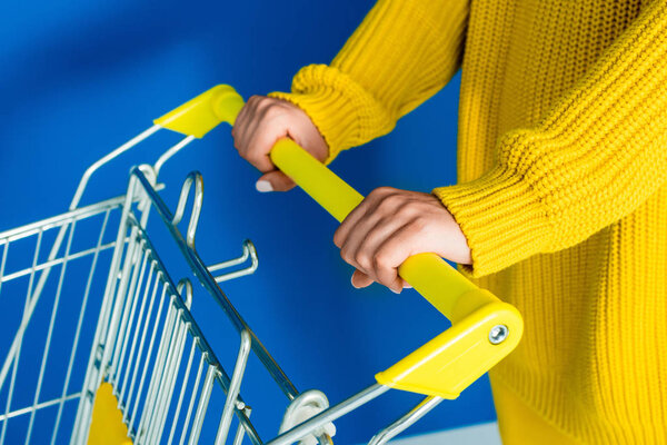 Cropped view of woman in yellow clothes holding shopping cart on blue background