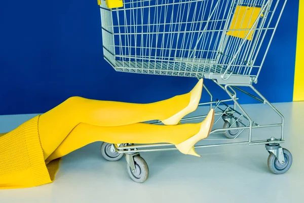 青色の背景のショッピング カートによって黄色の靴の女性の足のビューをトリミング  — 無料ストックフォト