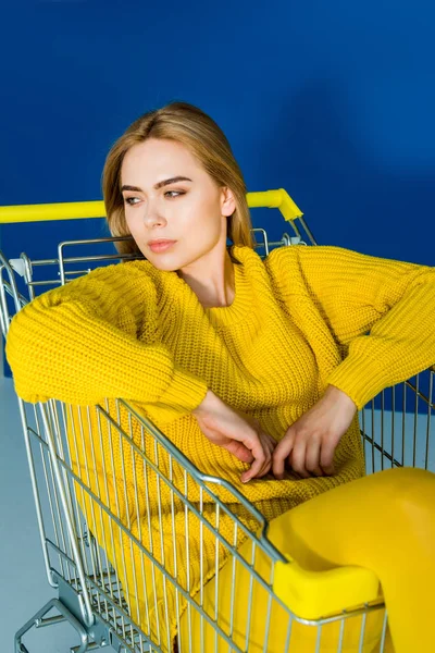 Female fashion model in yellow clothes sitting in shopping cart on blue background