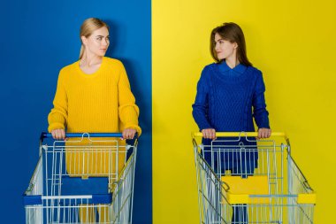 Smiling young women with shopping carts looking at each other on blue and yellow background