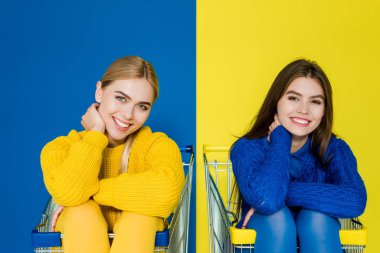 Attractive young girls having fun sitting in shopping carts isolated on blue and yellow background