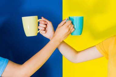 Cropped view of colorful cups in women hands on blue and yellow background clipart