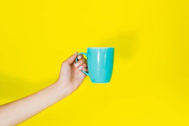 Cropped view of female hand with blue cup on yellow background clipart