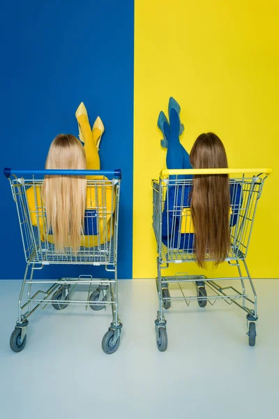Вид Сзади Женщин Моделей Сидящих Тележках Покупок Синем Желтом Фоне — стоковое фото