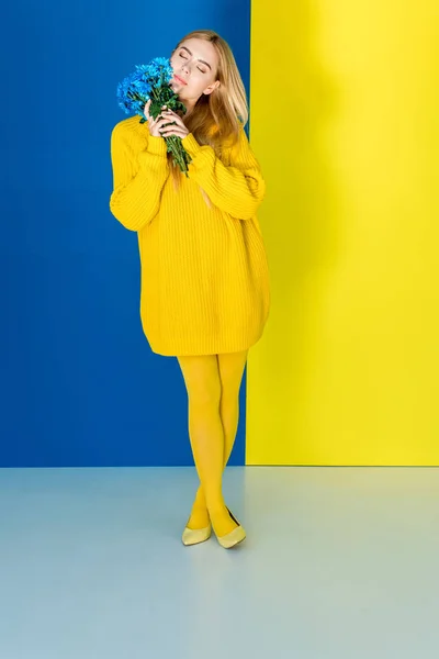 青と黄色の背景に青い花を保持している黄色の衣装で魅力的なブロンドの女性  — 無料ストックフォト