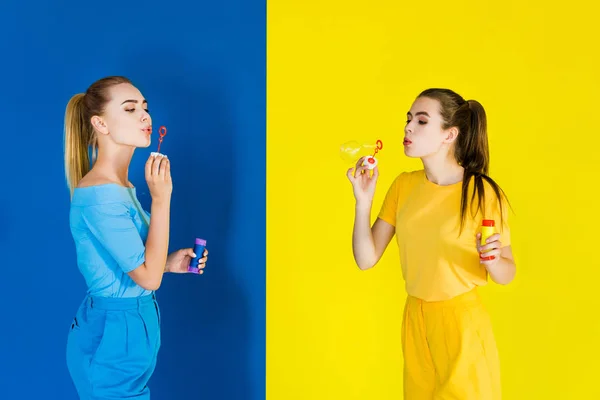 Beautiful Brunette Blonde Girls Blowing Bubbles Blue Yellow Background — Free Stock Photo