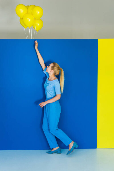 Beautiful blonde girl reaching out for yellow balloons on blue and yellow background