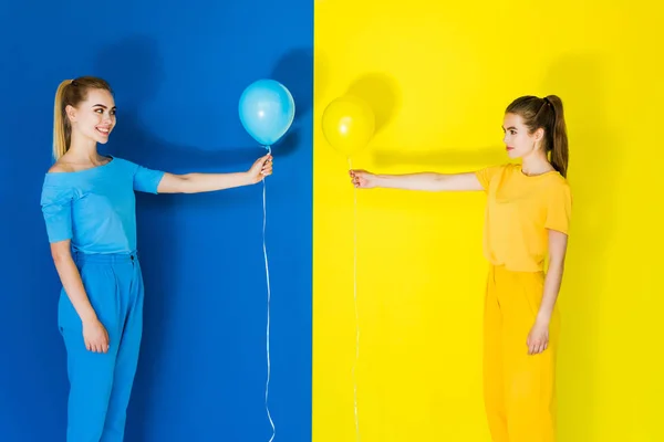 美丽的金发女郎和黑发女孩在蓝色和黄色的背景下互相拿着气球 — 图库照片