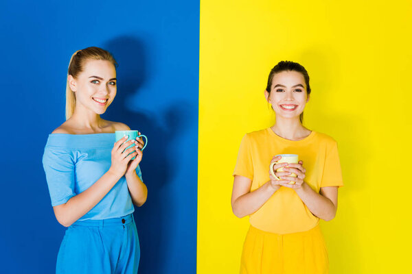 Smiling stylish women holding cups on blue and yellow background