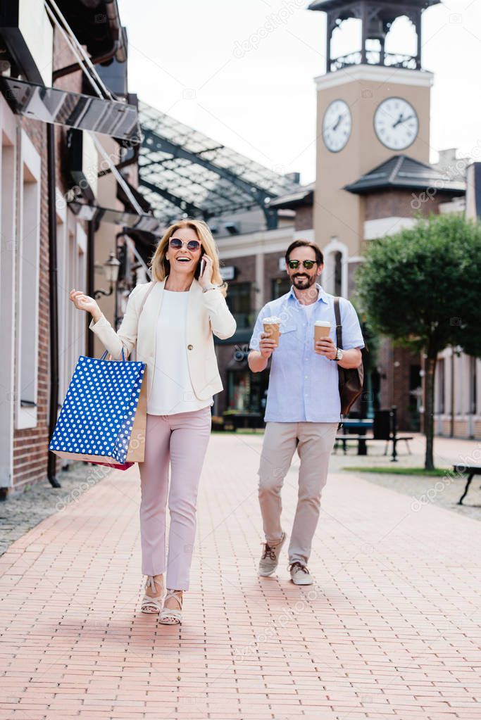 wife talking by smartphone and holding shopping bags, husband carrying disposable coffee cups on street