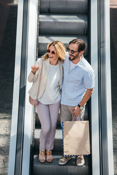 high angle view of happy couple in sunglasses looking away on escalator in shopping mall