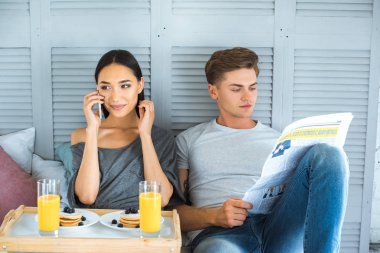 smartphone, gazete ve evde yatakta kahvaltı ile çok ırklı çift portresi