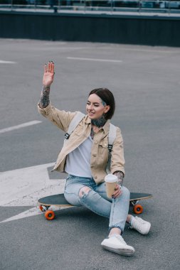 gülümseyen longboard kahve fincanı ile oturan ve el sallayarak dövmeli kadın 
