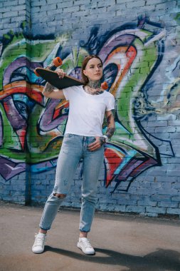 attractive girl with tattoos holding skateboard over shoulder near wall with graffiti clipart
