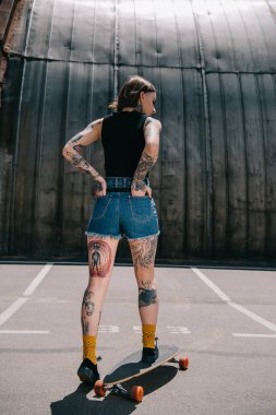 rear view of stylish tattooed girl with hands in pockets skateboarding at parking lot  clipart