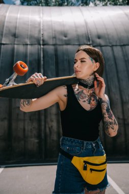 young stylish tattooed girl looking away and holding skateboard over shoulder  clipart