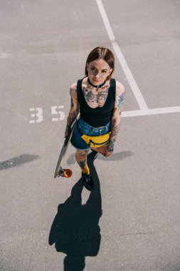high angle view of stylish tattooed girl holding skateboard and looking at camera at parking lot  clipart