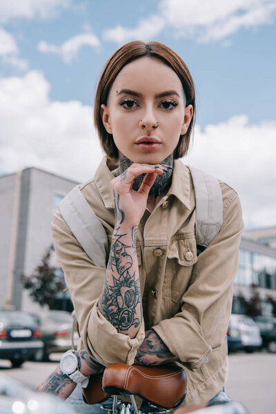 portrait of young tattooed woman looking at camera while sitting on bicycle at street 