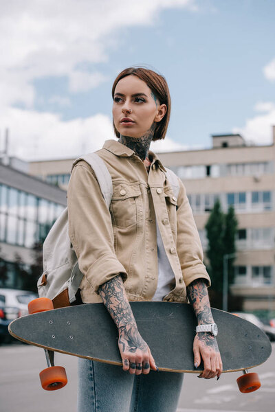 stylish tattooed woman looking away and holding longboard at street