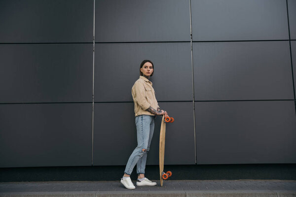 stylish tattooed woman standing with skateboard against black wall 