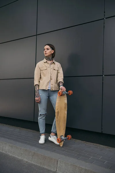 young tattooed woman standing with skateboard against black wall